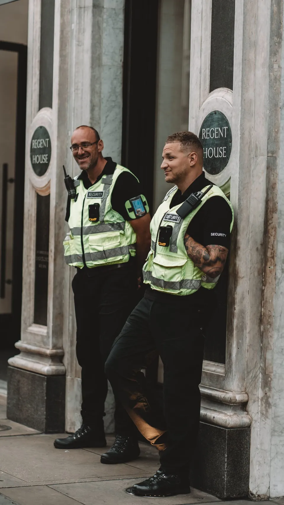 Deux agents de sécurité en gilet haute visibilité, souriants, à l’entrée d’un immeuble